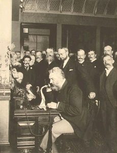 Figure 1: This image depicts Alexander Graham Bell with his invention, the telephone, surrounded by a room full of men.